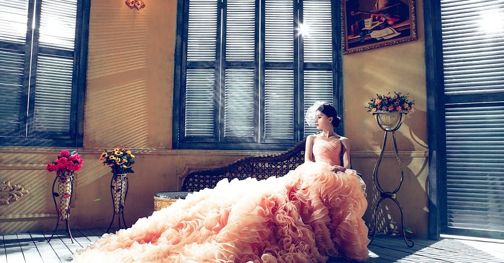 A bride in a luxurious gown sits gracefully in a sunlit room, exuding elegance.